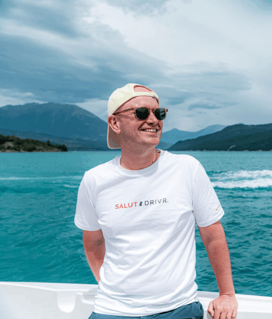 Gründer von Sportwagen Erlebnisreisen (Ernst Behrens) auf einem Boot vor malerischer Bergkulisse mit einem Salut Drivr T-Shirt.