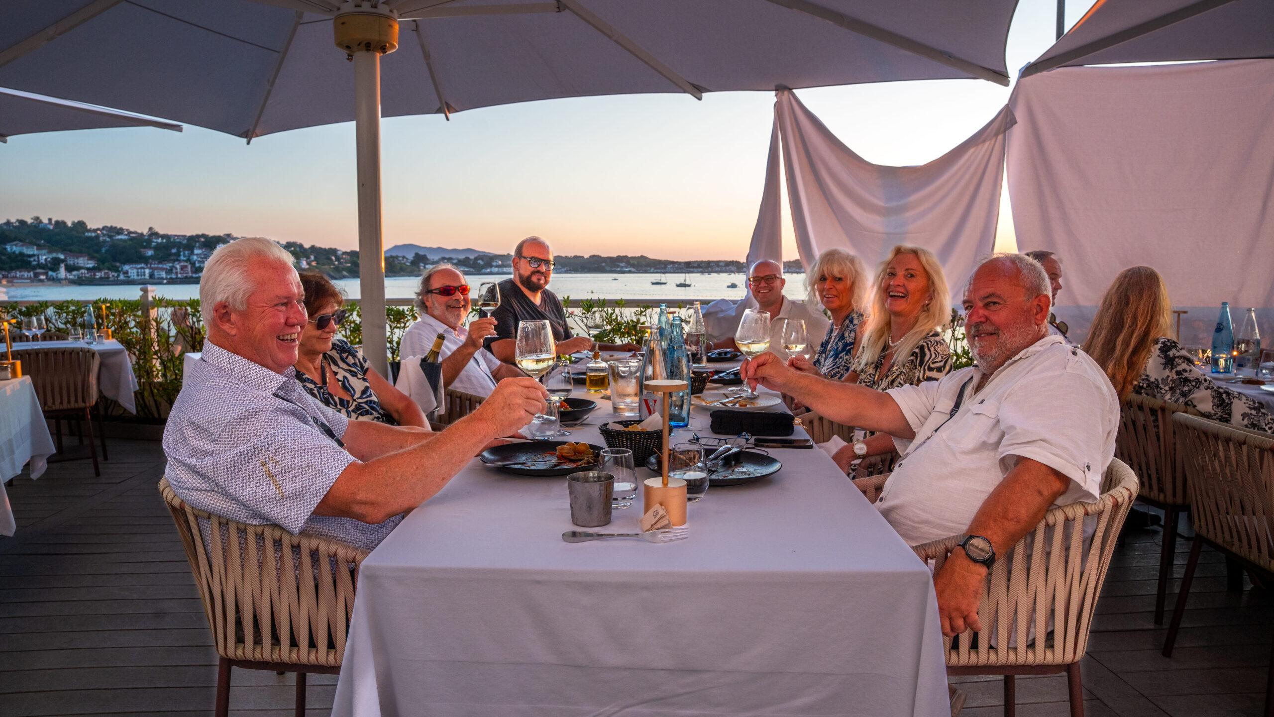 Teilnehmer einer Sportwagen Erlebnisreise stoßen beim Abendessen am Meer vor Sonnenuntergang an.