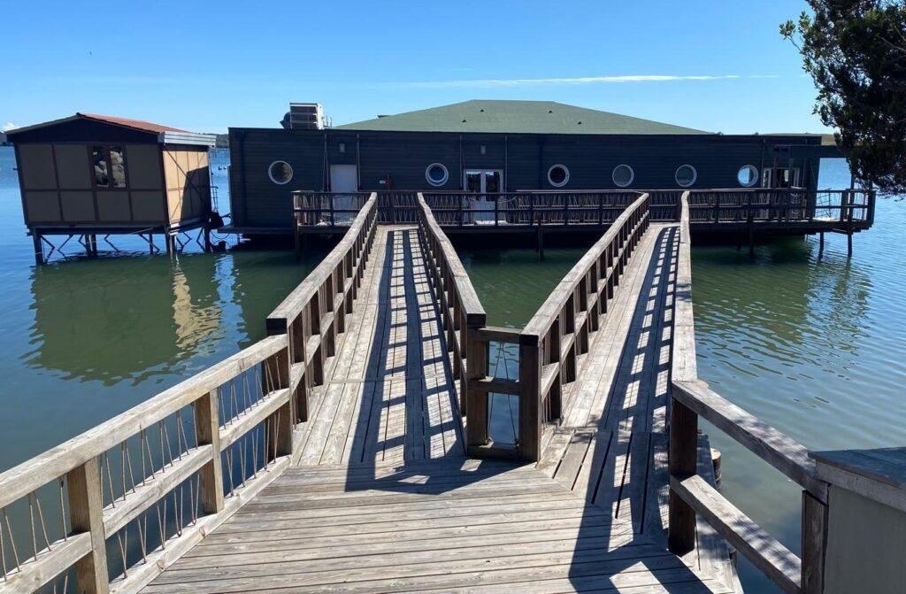 Holzsteg führt zu schwimmendem Gebäude auf ruhigem See unter blauem Himmel.