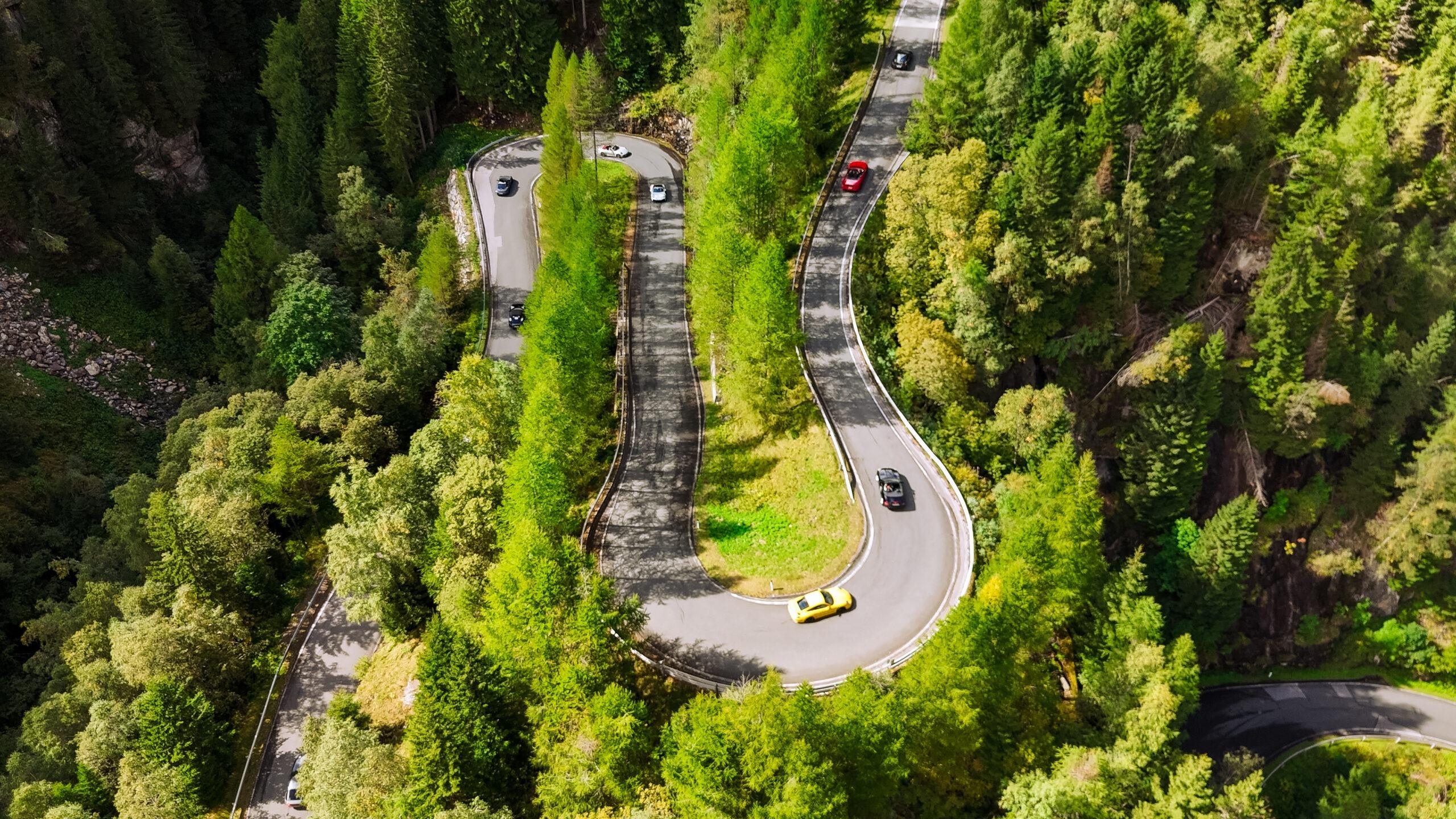 Supercars fahren durch malerische Kurvenstraßen in den Alpen, umgeben von dichtem Wald.