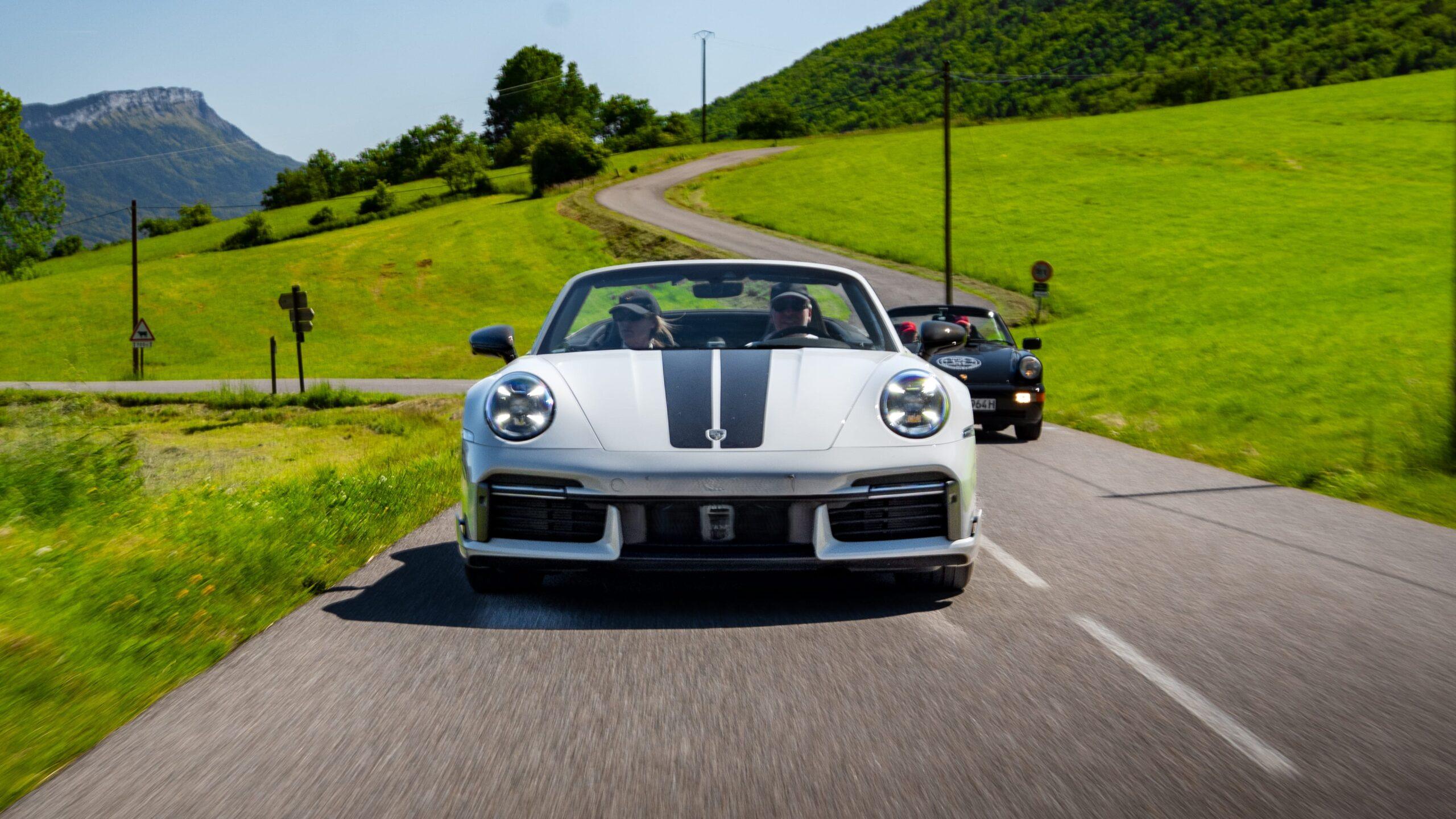 Weiße Sportwagen fahren auf malerischer Straße durch grüne Landschaften in den Alpen.