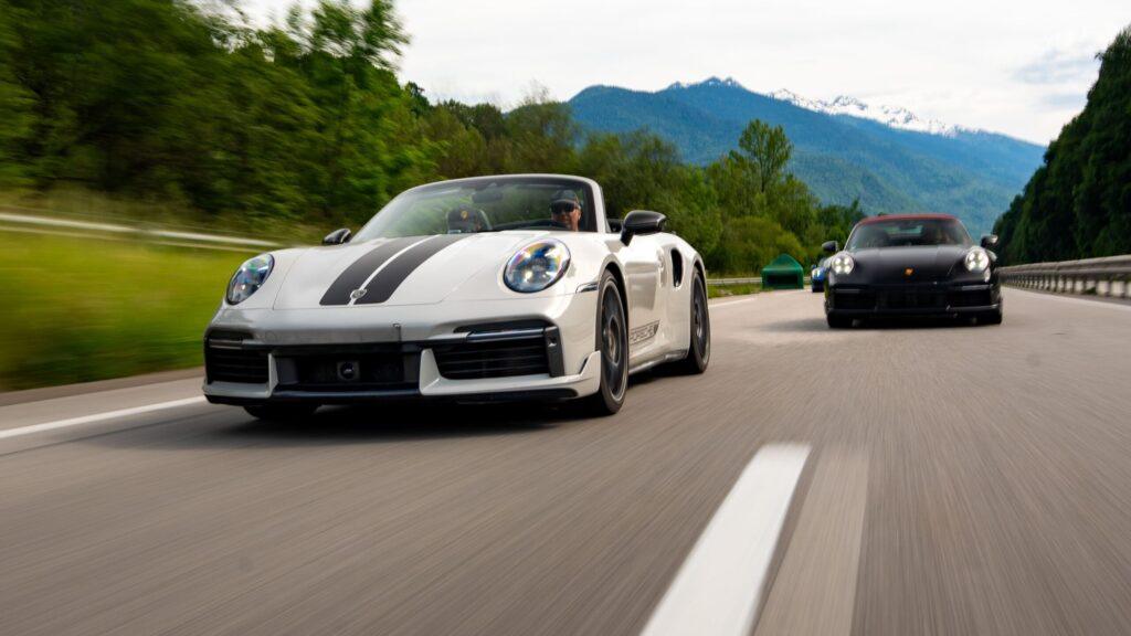 Zwei Sportwagen gleiten elegant entlang einer Bergstraße, umgeben von grünen Wäldern und alpiner Kulisse.
