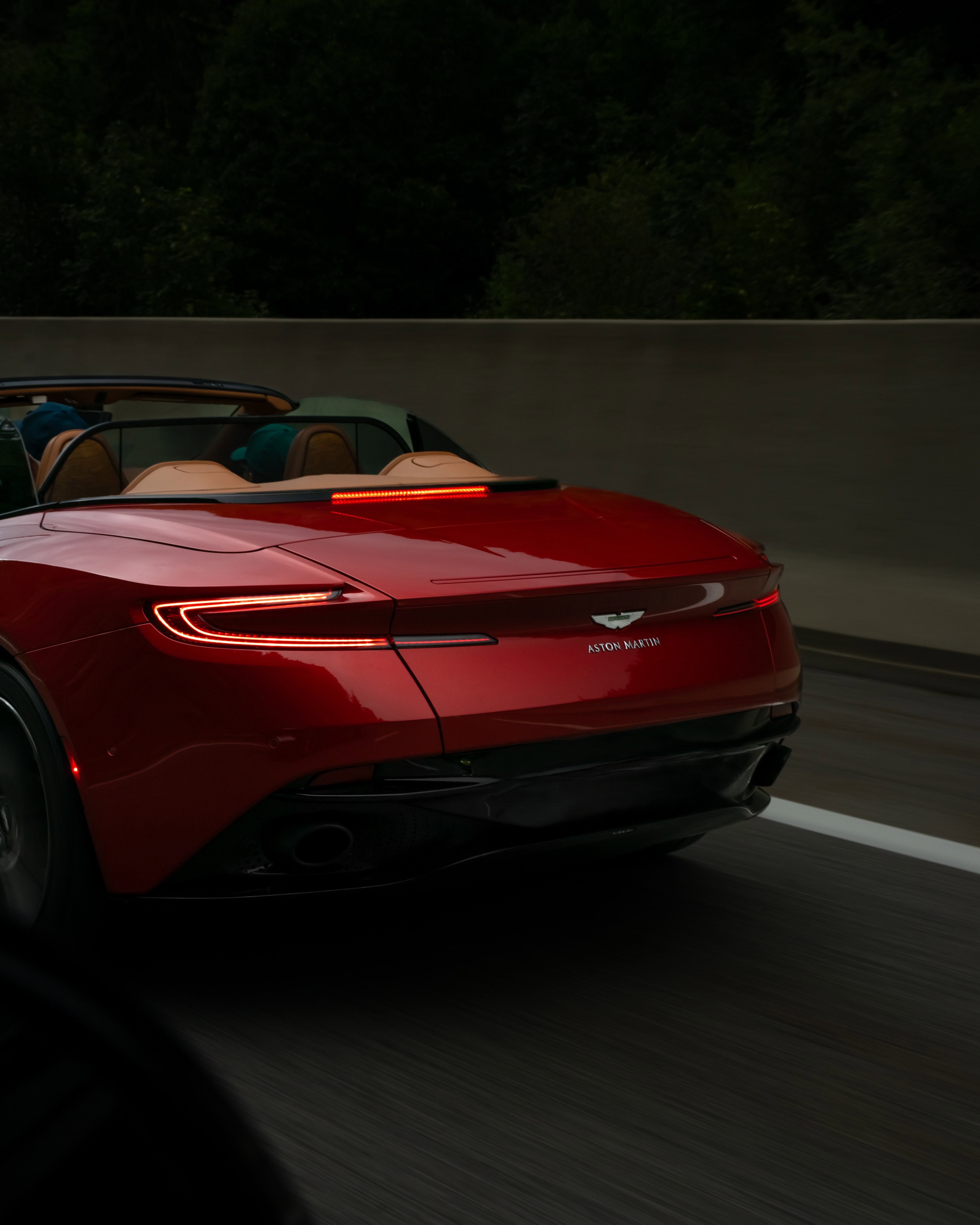 Ein roter Aston Martin Cabrio fährt dynamisch auf einer malerischen Straße, umgeben von grünen Bäumen.