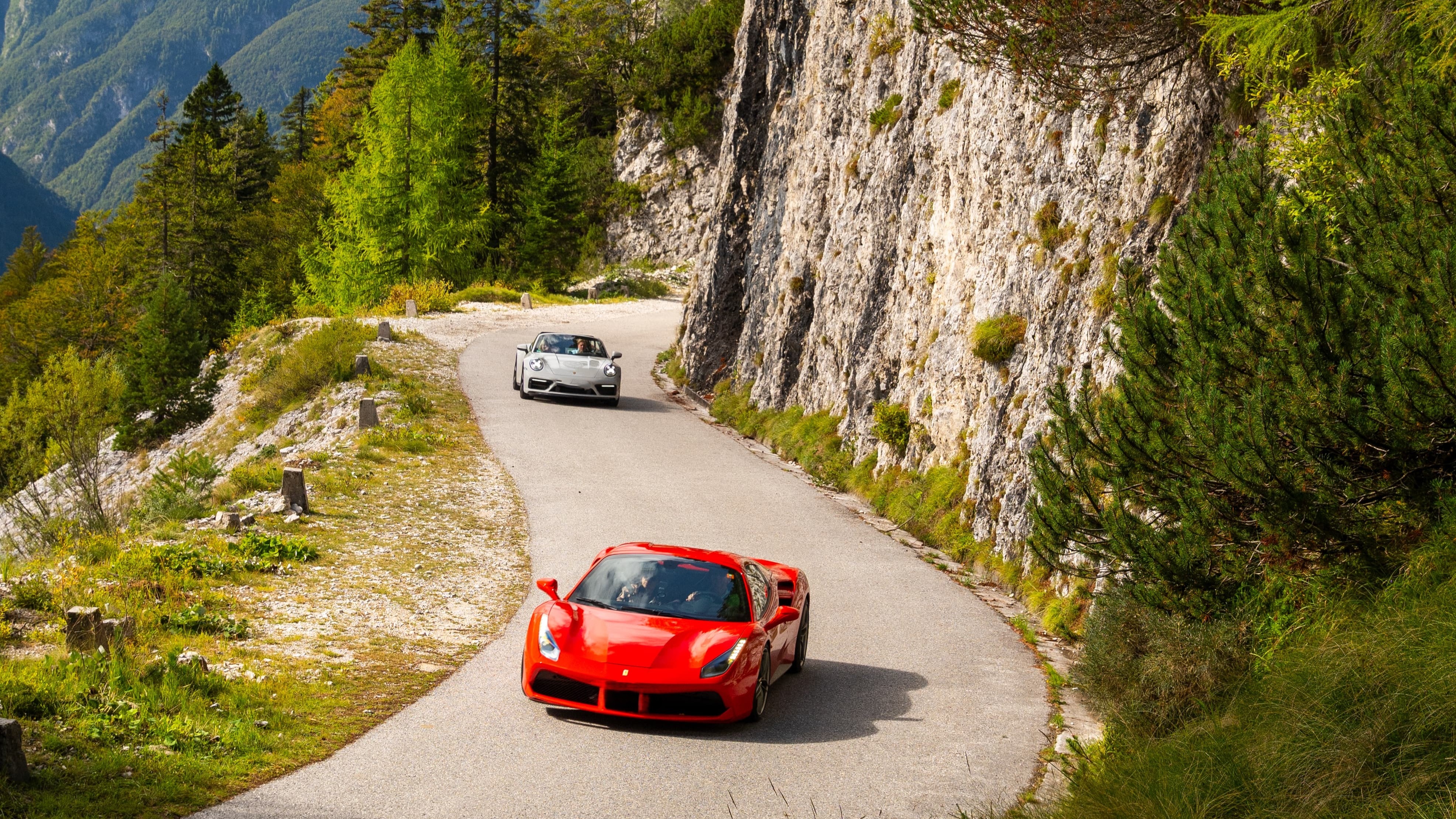 Rote und silberne Sportwagen fahren auf kurvenreicher Bergstraße in den Alpen.