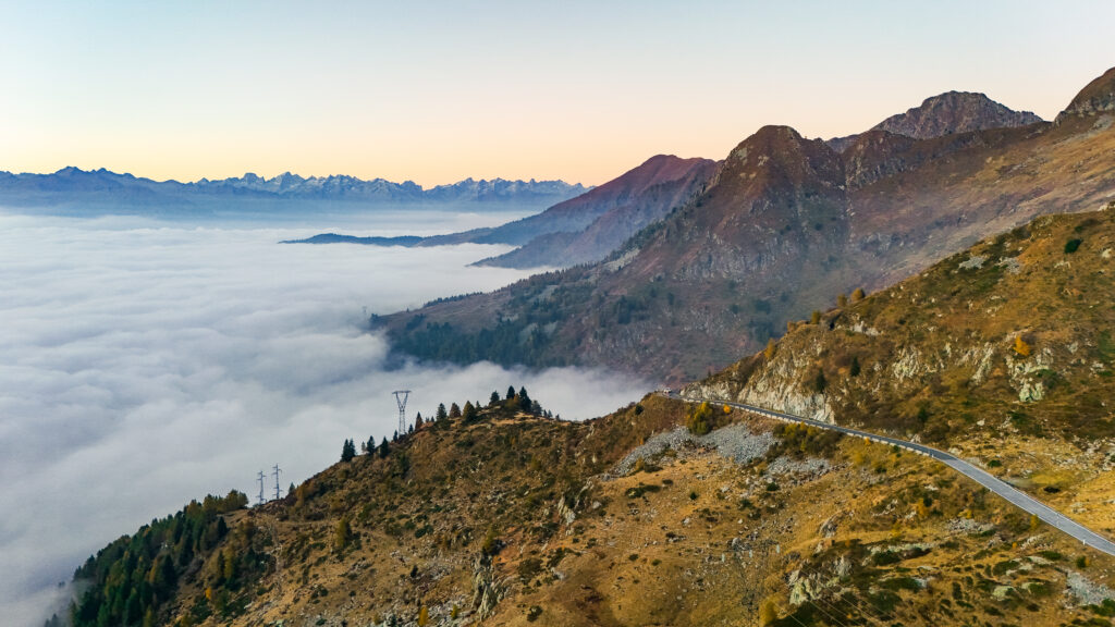 Kurvenreiche Bergstraße durch die Alpen bei Sonnenaufgang, umgeben von Wolkenmeer.