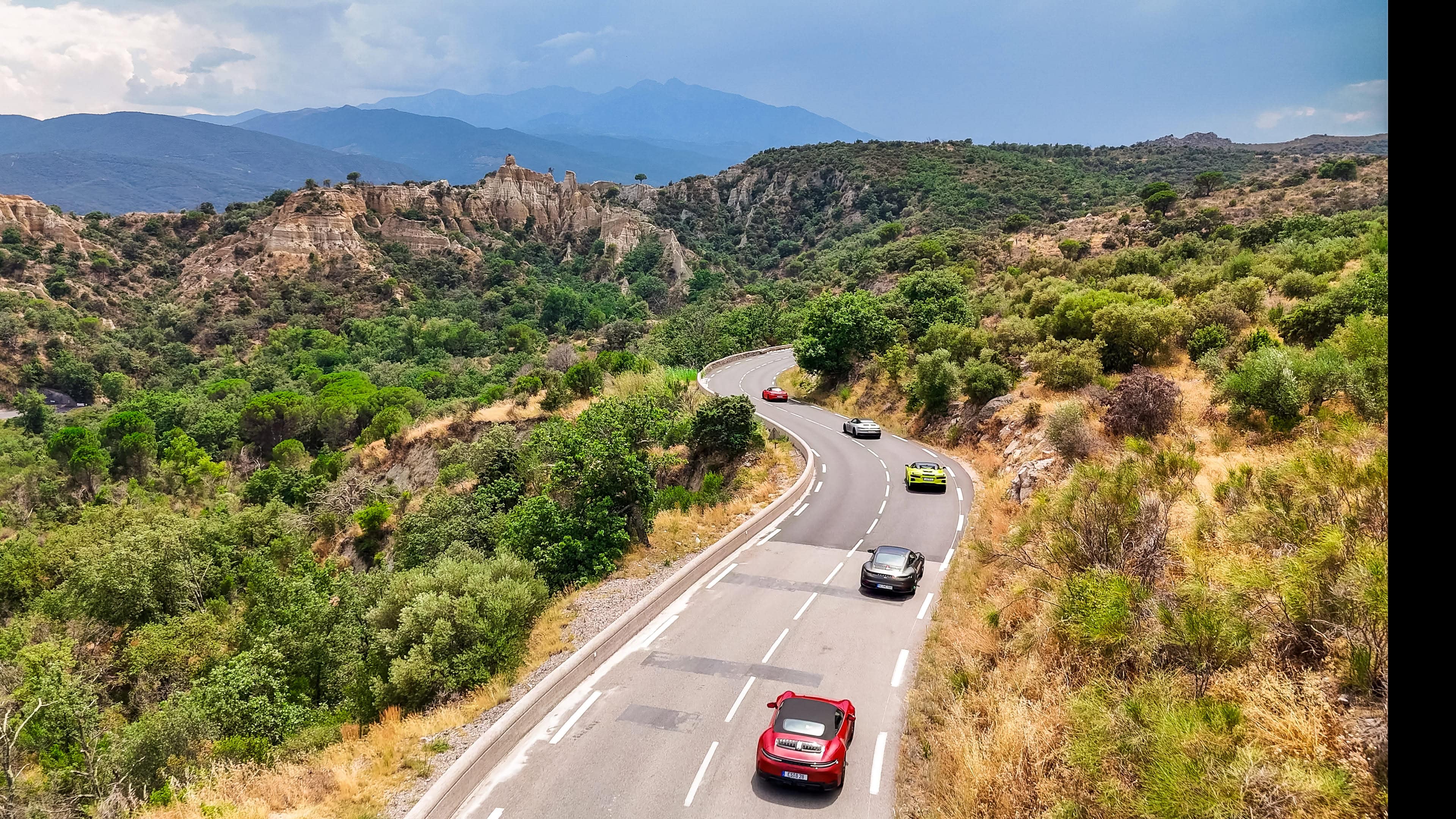 Kurvenreiche Straße durch grüne Hügel, umgeben von majestätischen Bergen und sportlichen Autos. Abenteuer pur!