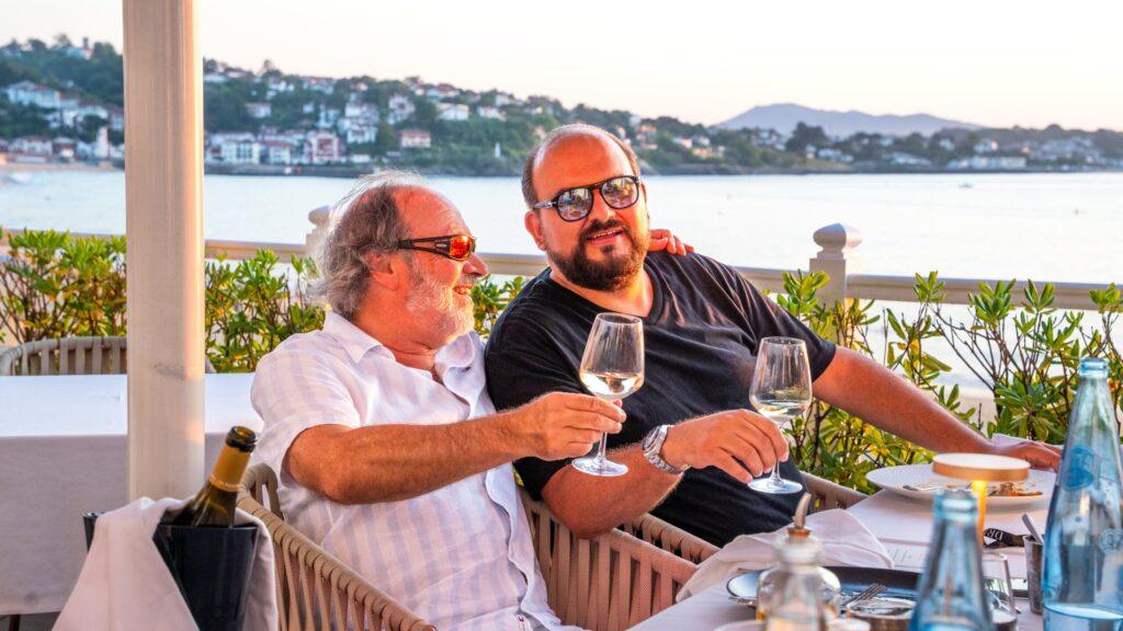 Zwei Herren stoßen an Gläsern in einem eleganten Restaurant mit Blick auf das Wasser und die Küste im Sonnenuntergang.