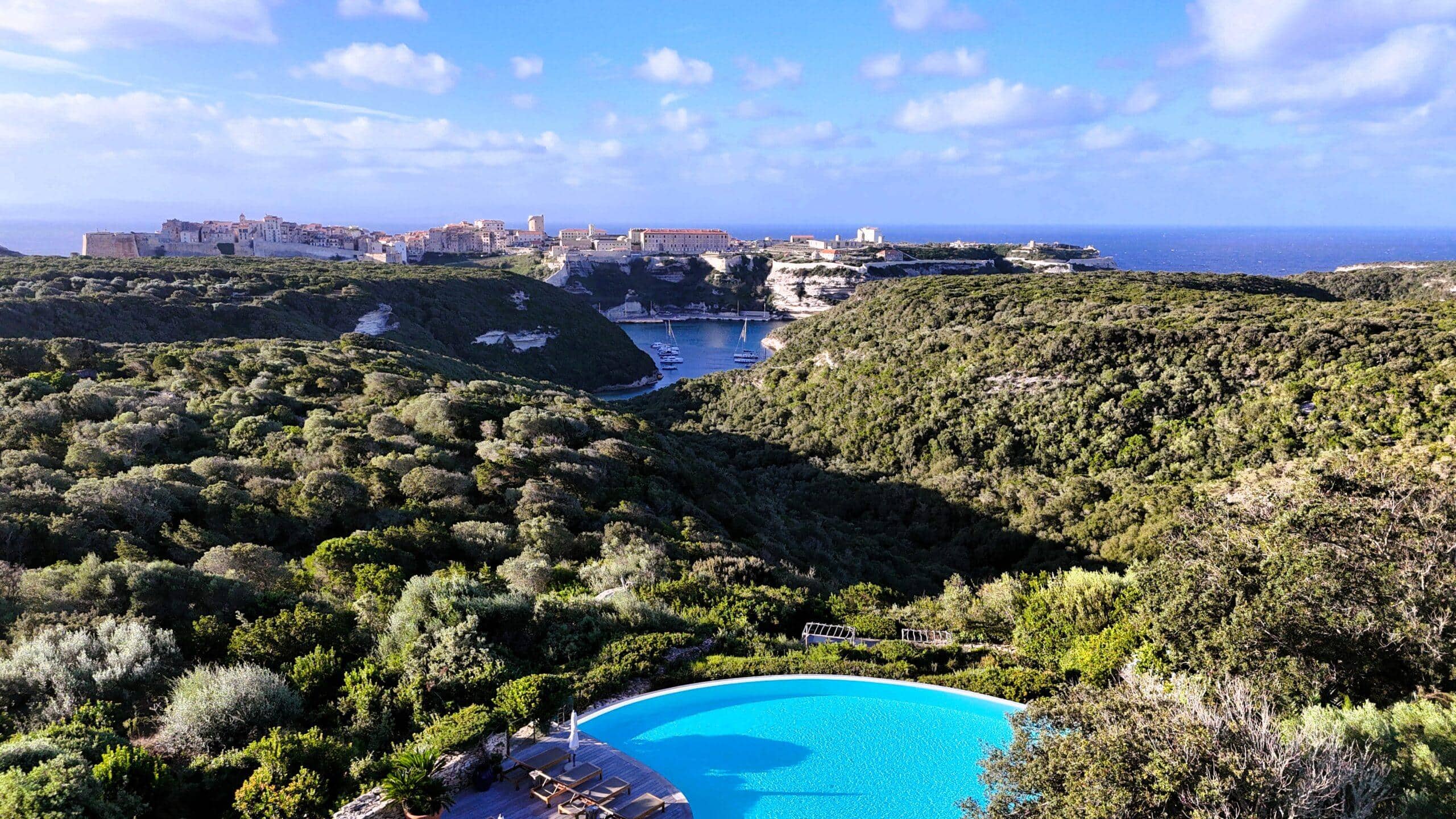 Panoramablick auf die Küste von Korsika mit azurblauem Pool und malerischer Altstadt im Hintergrund.