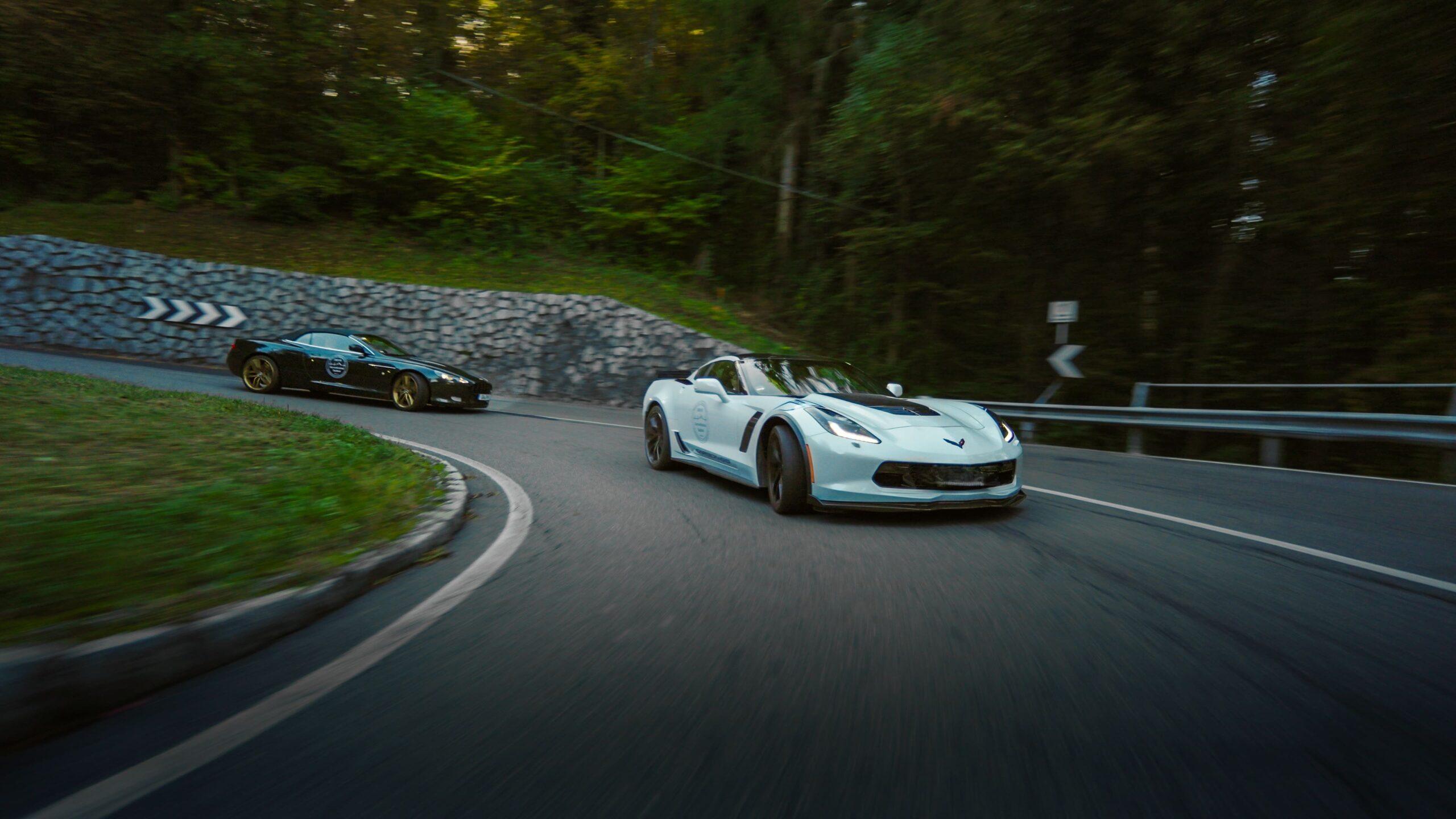 Zwei Sportwagen fahren dynamisch auf einer kurvenreichen Bergstraße im Wald.