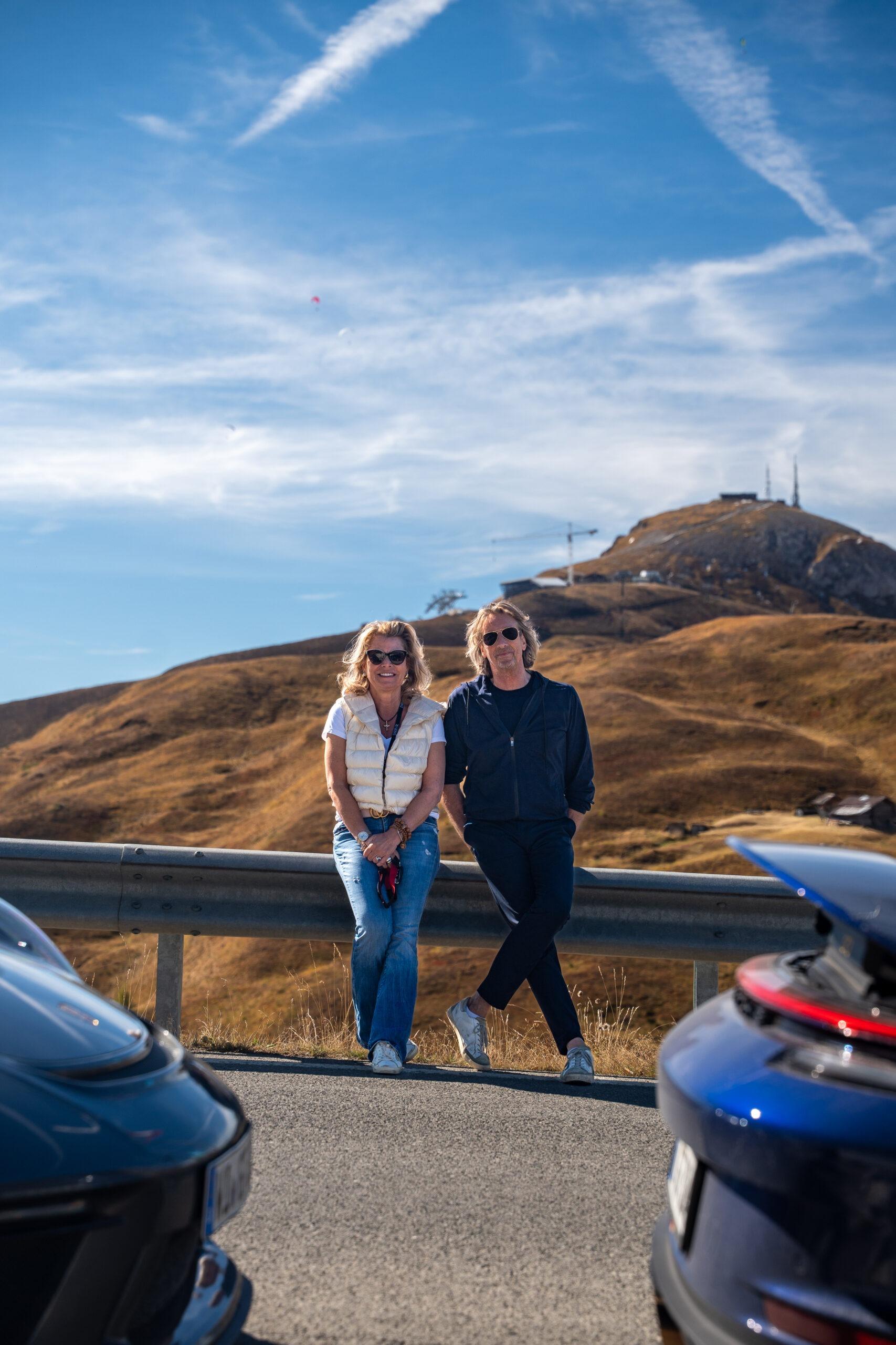 Paar posiert entspannt neben einem Porsche an einer Passstraße während der Sportwagen Erlebnisreise in Südtirol.