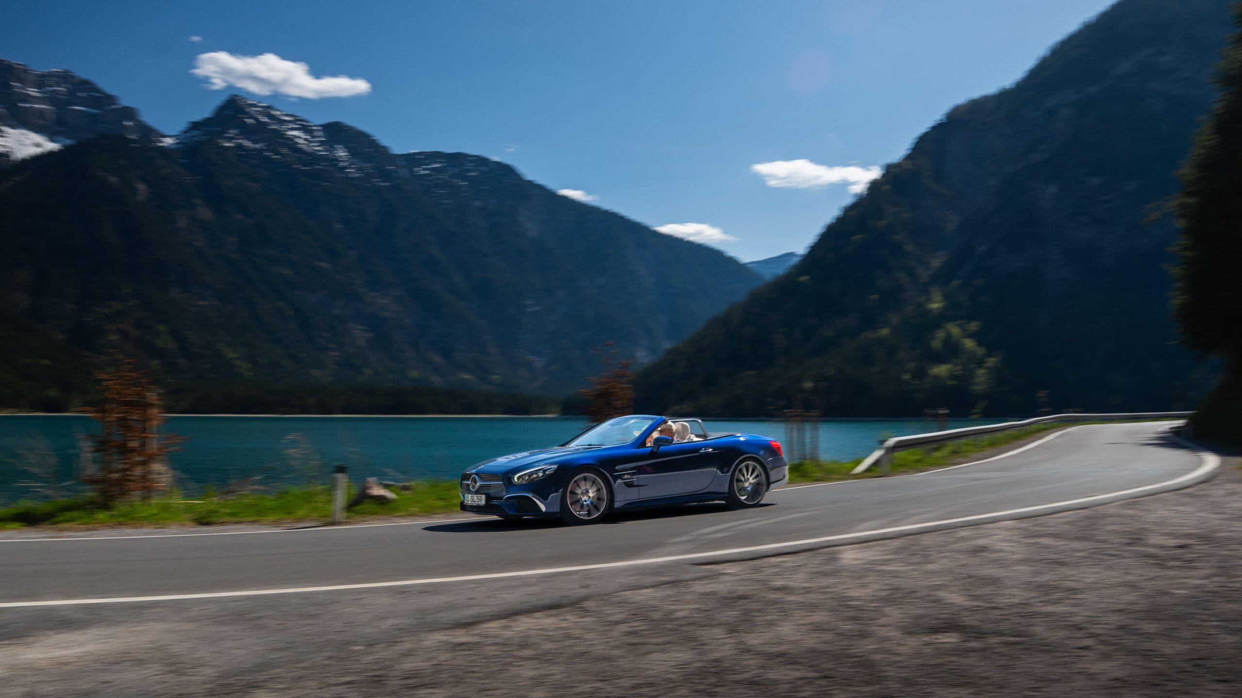 Ein blauer Sportwagen fährt entlang eines malerischen Sees in den Alpen.
