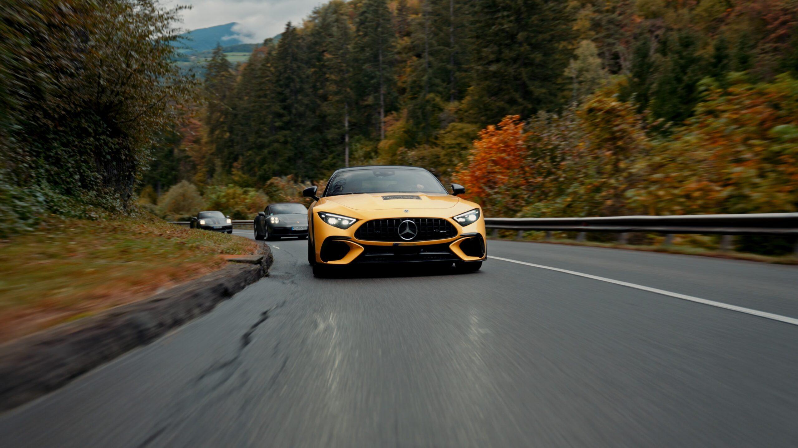 Gelber Sportwagen fährt entlang einer malerischen Straße, umgeben von bunten Herbstbäumen in den Alpen.