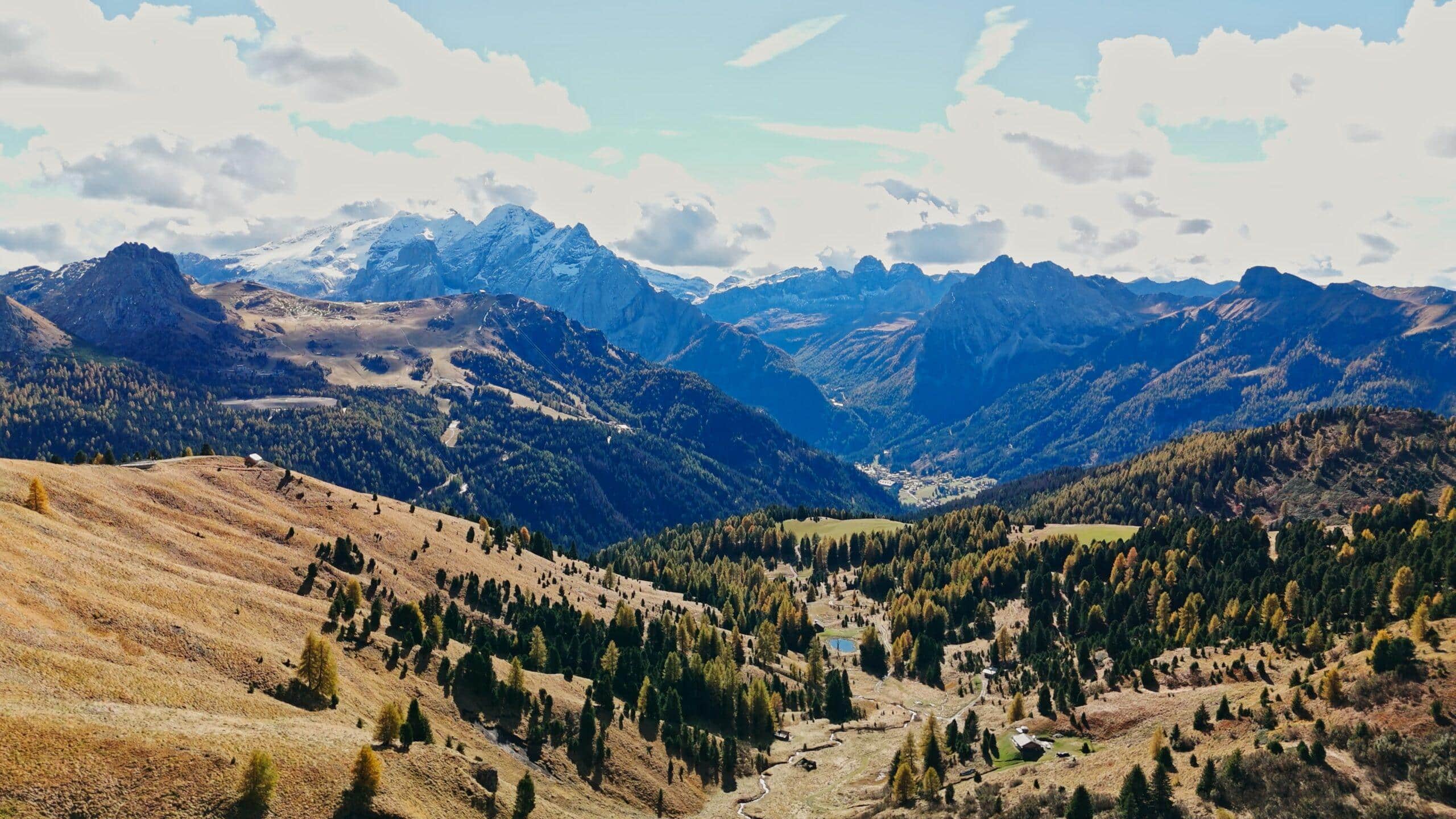 Majestätische Alpenlandschaft mit sanften Hügeln, bunten Wäldern und beeindruckenden Gipfeln unter strahlend blauem Himmel.