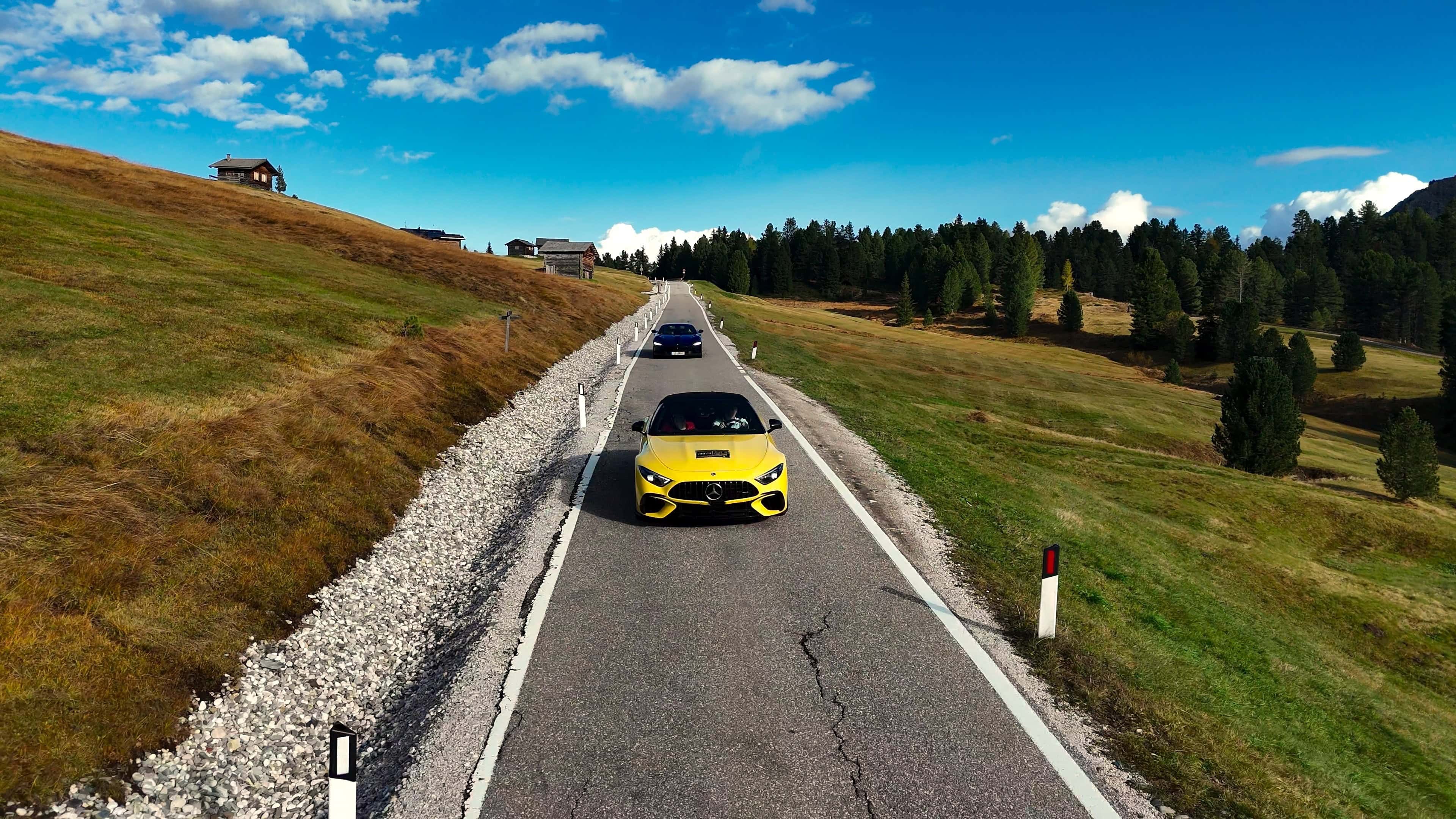 Ein gelber Sportwagen fährt auf einer malerischen Gebirgsstraße in den Alpen. Grüne Wiesen und blauer Himmel schaffen eine traumhafte Kulisse.