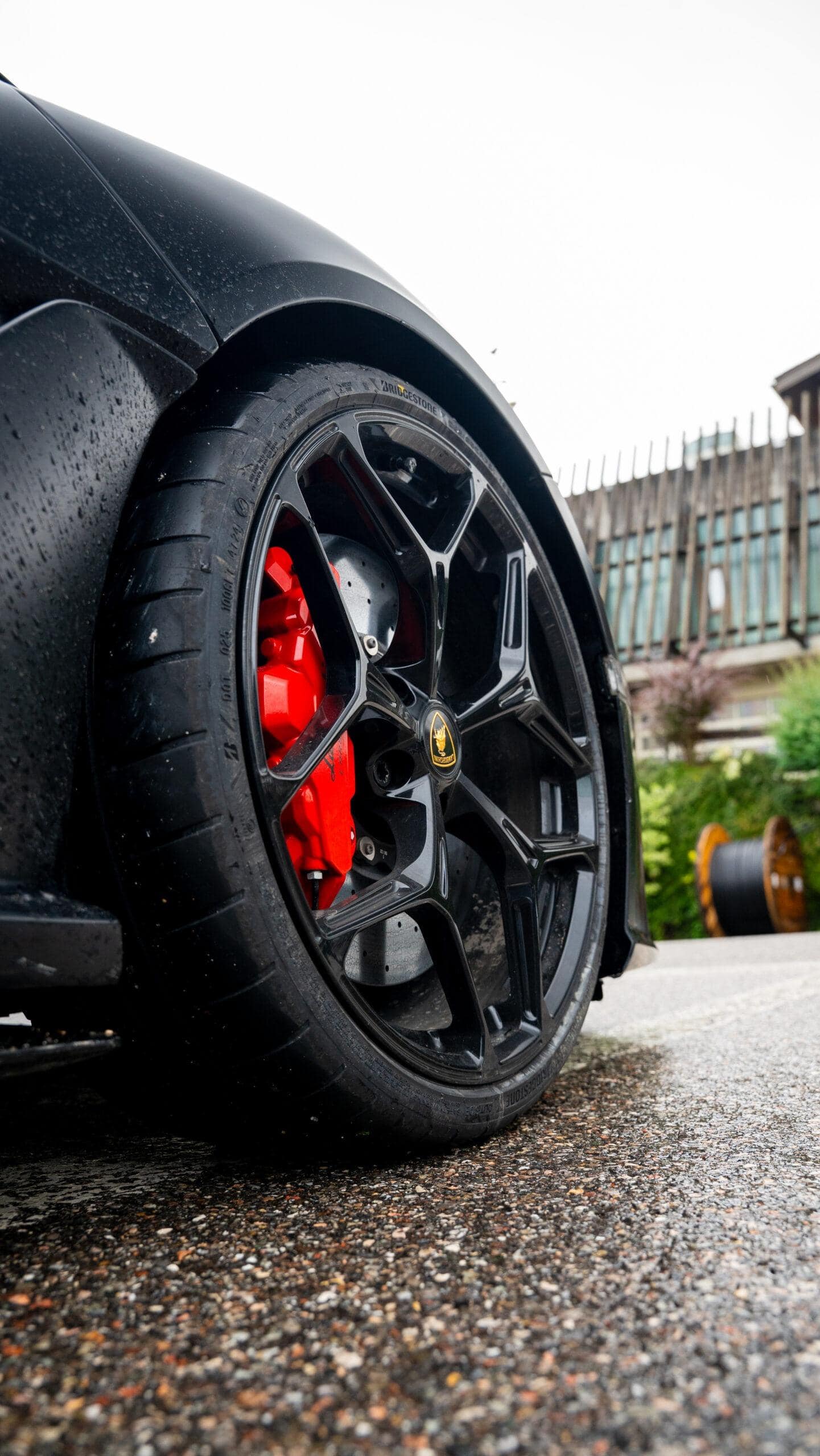 Schwarzes Sportwagenrad mit leuchtend roten Bremssätteln auf regennasser Straße, luxuriöse Atmosphäre im Hintergrund.