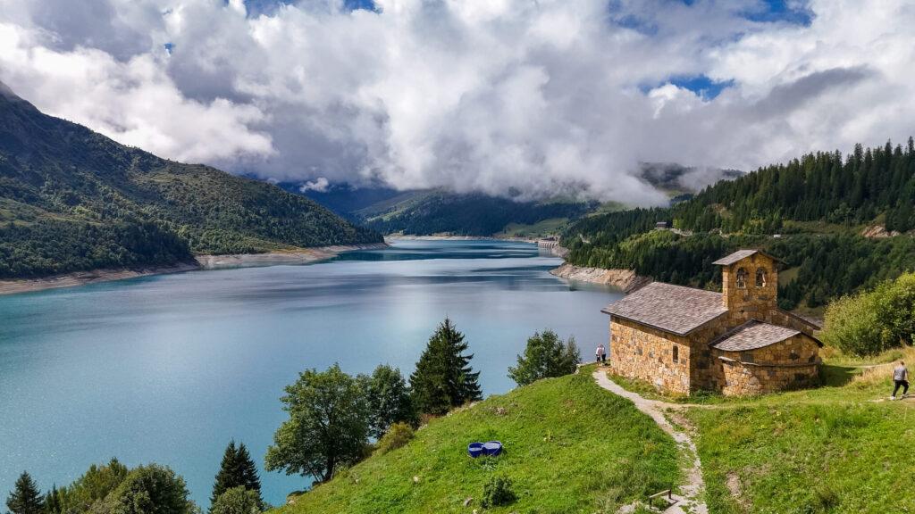 Idyllische Berglandschaft mit einer malerischen Kapelle am glitzernden See, umgeben von grünen Hügeln.