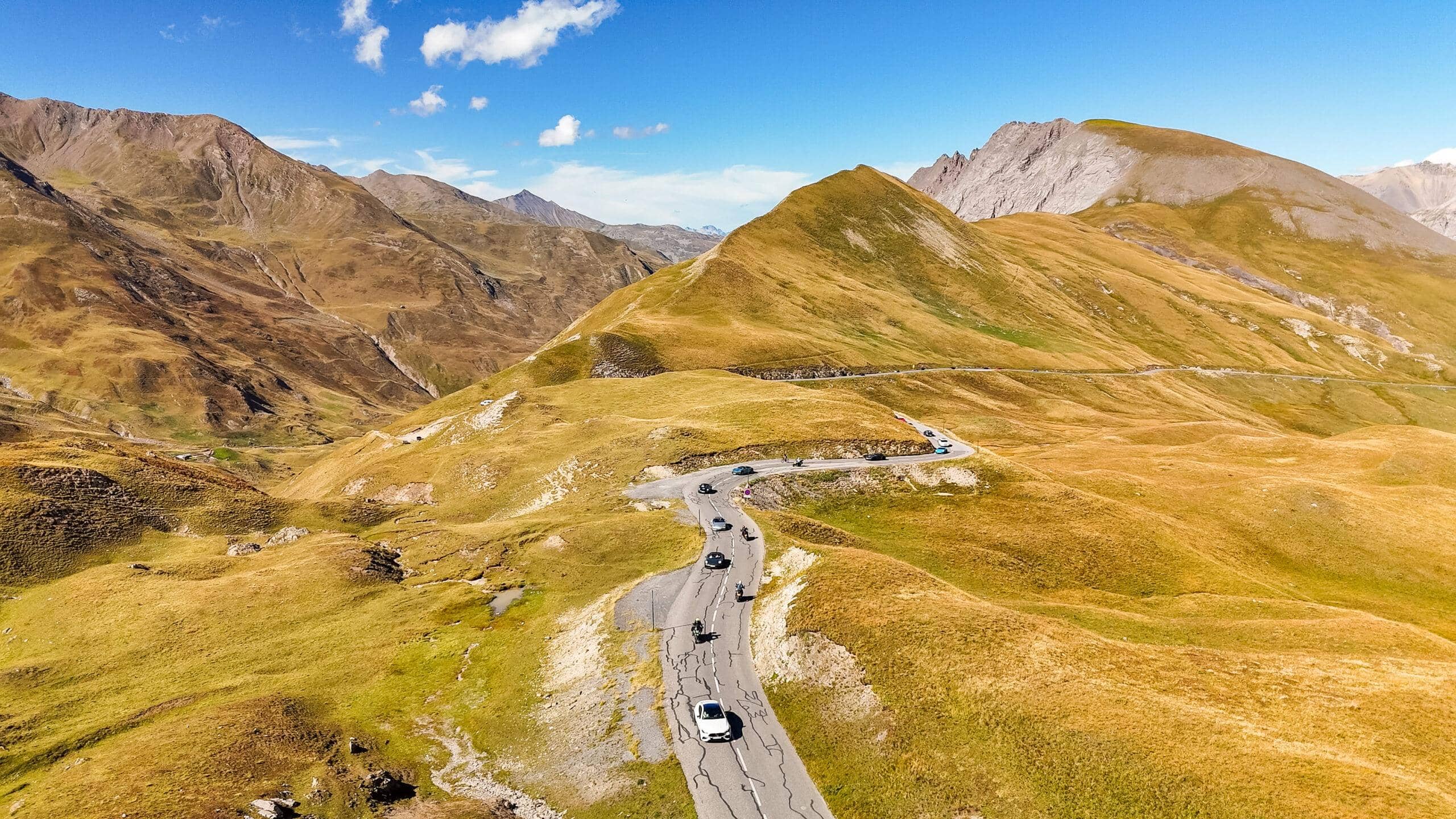 Kurvenreiche Straße zwischen majestätischen Bergen, umgeben von goldenen Wiesen und strahlend blauem Himmel.