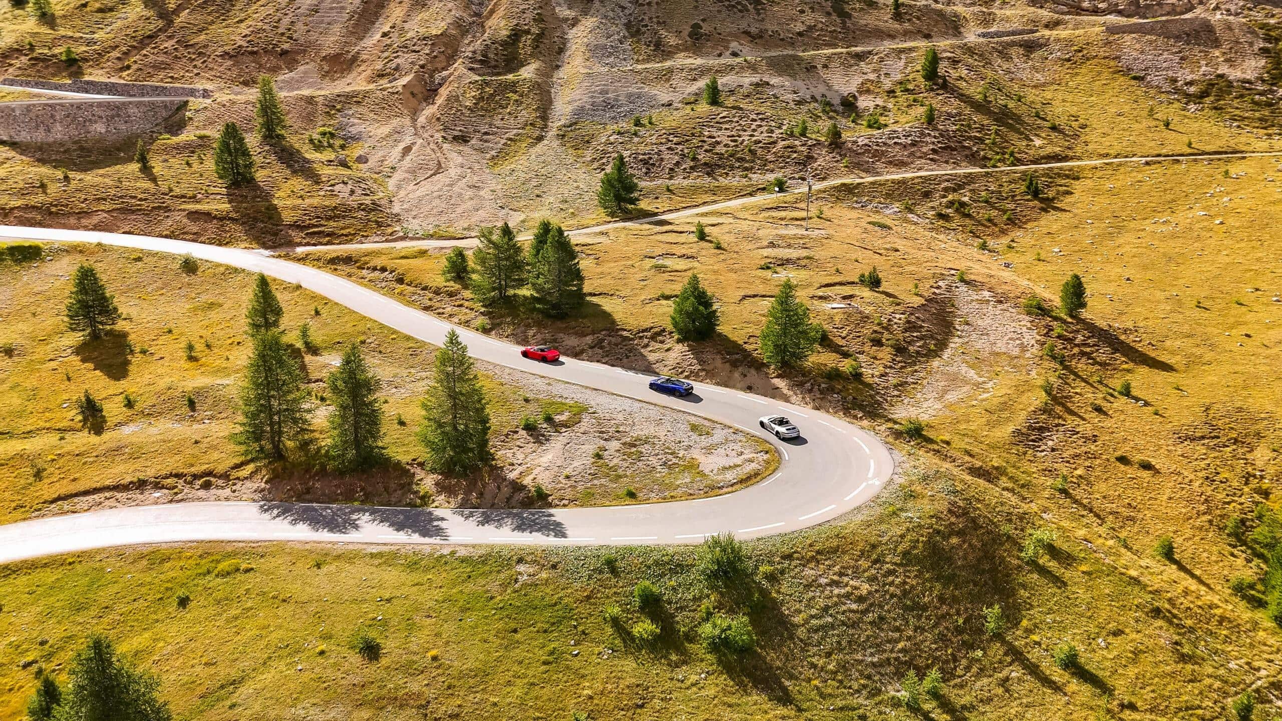 Drei Sportwagen kurven durch die malerischen Alpenlandschaften unter strahlend blauem Himmel.