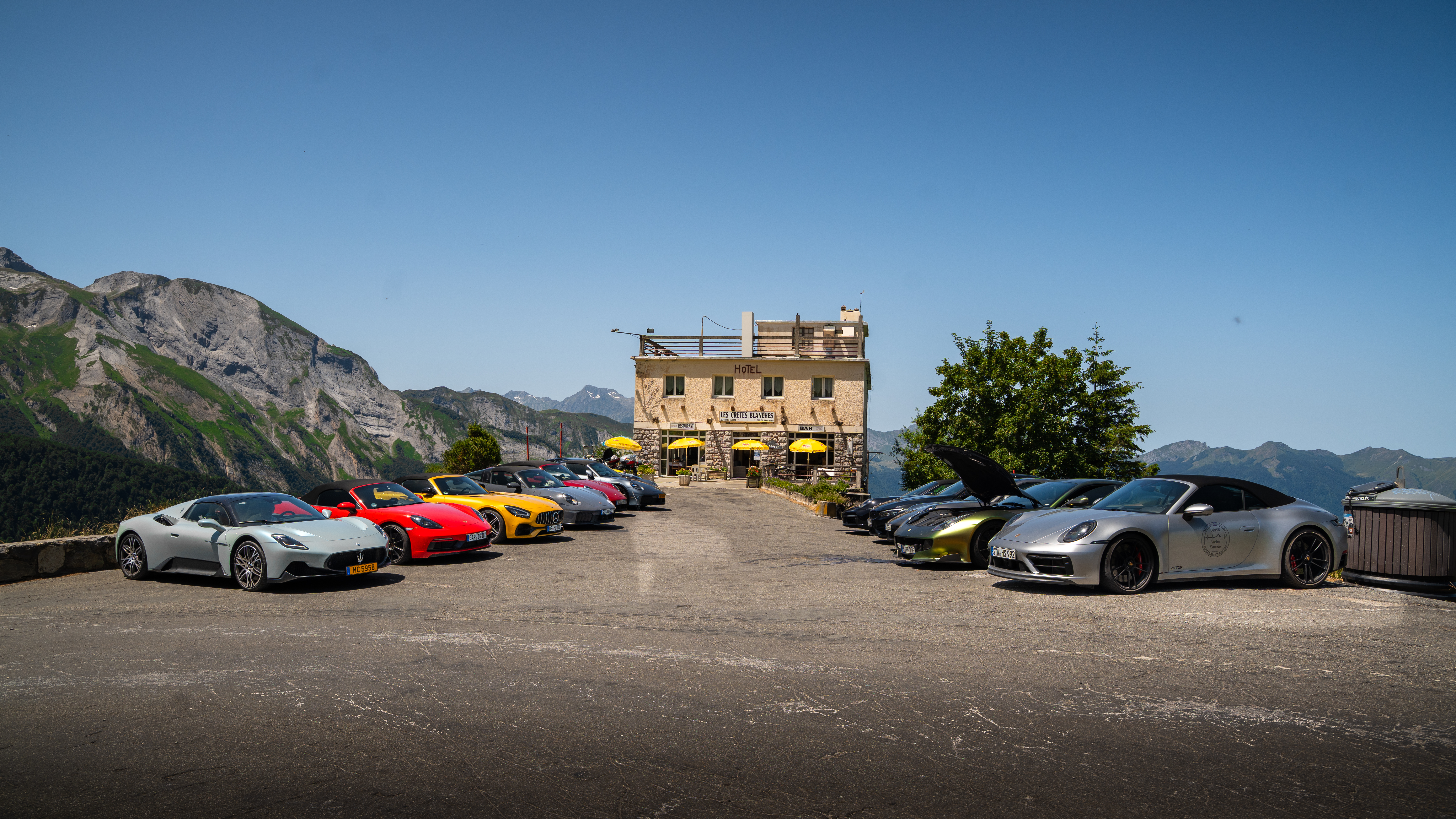 Eine Gruppe von Sportwagen parkt vor einem Alpenpanorama und einem Berghotel.
