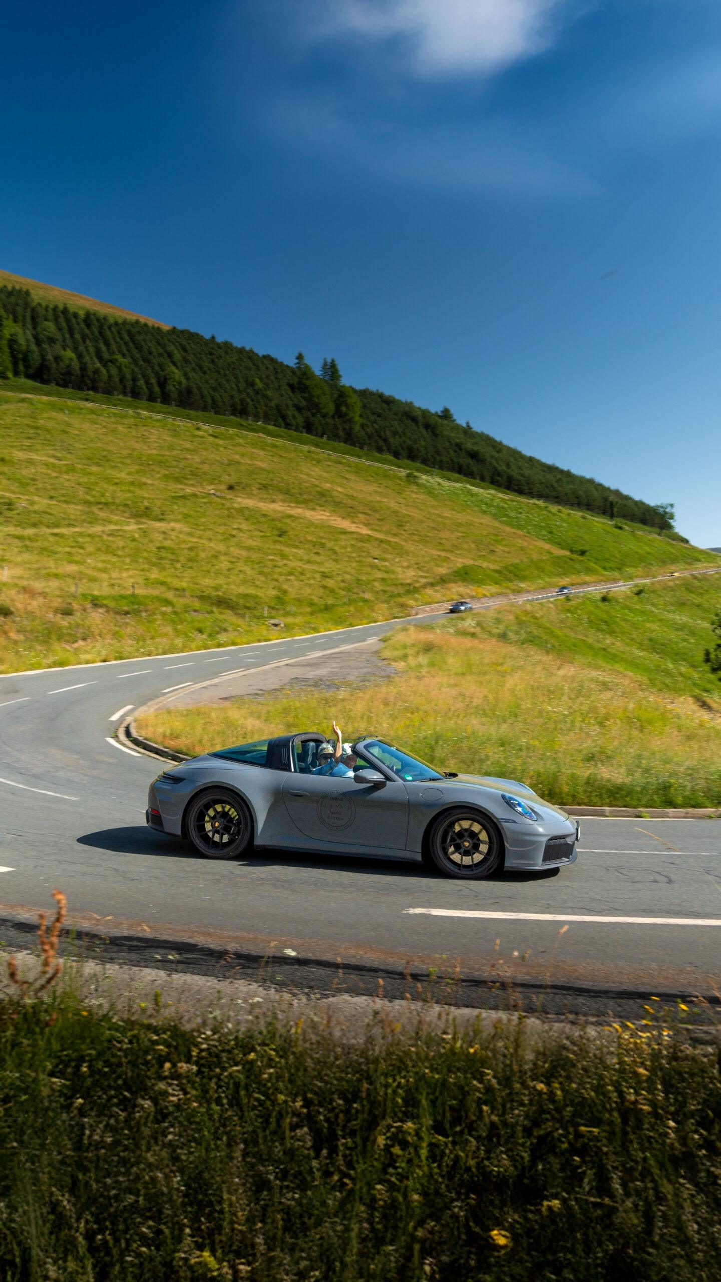 Grauer Porsche 911 Targa mit winkendem Beifahrer auf einer geführten Sportwagen-Reise durch grüne Landschaft.