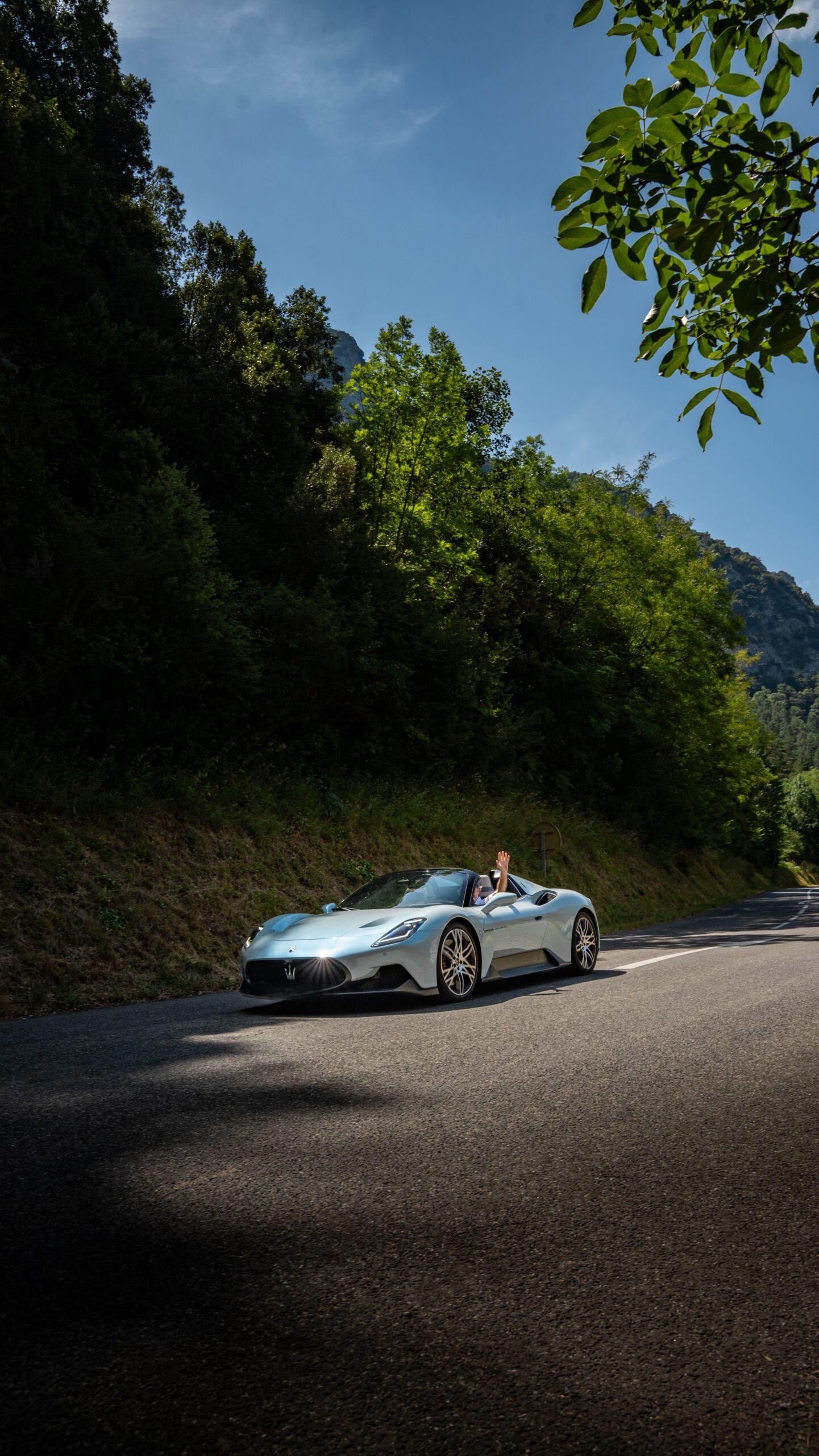 Sportwagen Erlebnisreisen: Fahrer im Maserati MC20 Cielo Cabrio grüßt auf sonniger Küstenstraße.
