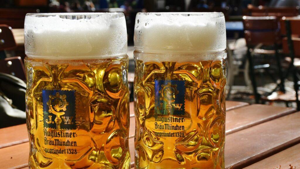 Zwei Krüge mit Bier auf einem Holztisch in einem sonnigen Biergarten.