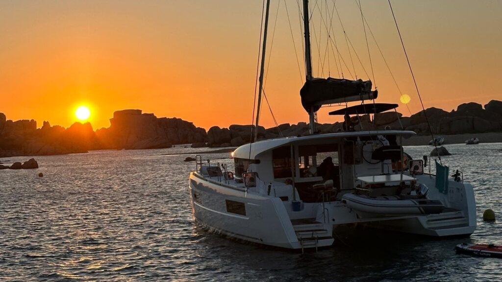 Ein Katamaran segelt bei Sonnenuntergang entlang einer felsigen Küstenlandschaft.