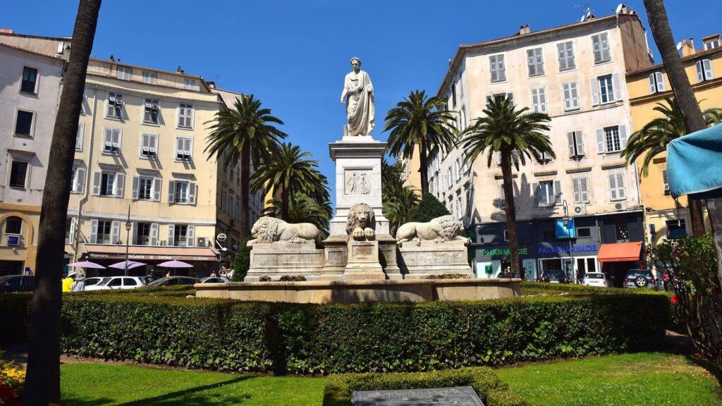 Elegante Statue umgeben von Palmen, idyllischer Platz mit lebhaftem Stadtleben in sonniger Atmosphäre.