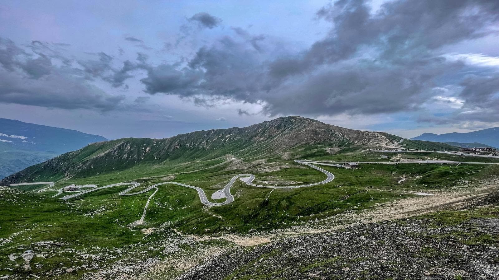 Kurvenreiche alpine Straße umgeben von grünen Wiesen und majestätischen Bergen, unter einem dramatischen Himmel.